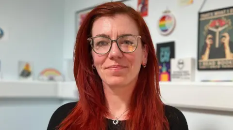 Kaye has long red hair and glasses. She's wearing a black top and a lanyard, seated indoors against a light blue wall decorated with framed posters and artwork