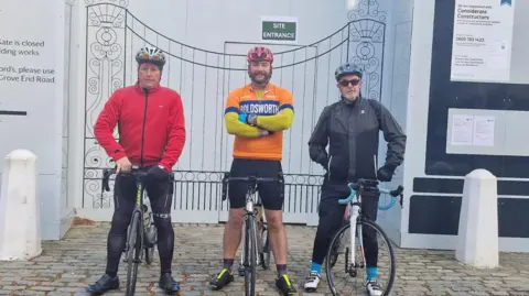 Goatacre Cricket Club Three cyclists stood with bikes and in sports gear on cobbles.