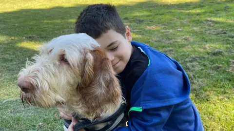 CHARLOTTE VOWDEN/BBC Ten-year-old Isaac Thompson, who as a a dark short back and side hair style and is wearing a blue hoodie with bright green piping down the zip line, hugs Hero from behind