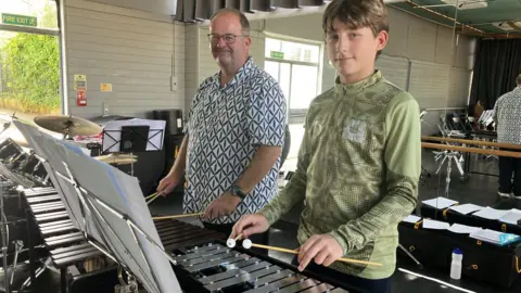 A young boy is standing in the front of the picture holding two beaters poised above a glockenspiel. An older man is standing in the background with his beaters on an xylophone. 