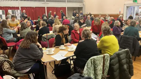 a lot of people sitting around trestle tables with cups of tea 