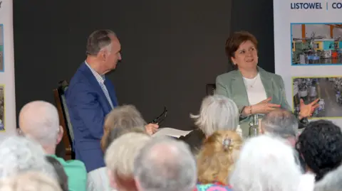 PA Media Nicola Sturgeon wearing a pale green suit over a white top, sitting on a stage next to author Andrew O'Hagan, wearing a blue suit.