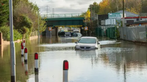 Cars, covered by water up to their lights, sit in flood waters on a road also obscured by flooding.