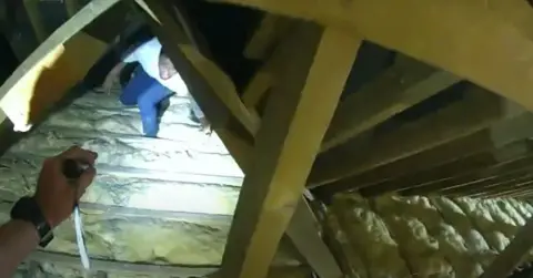 Essex Police A man walks across the rafters inside a house's loft towards someone in the foreground holding a torch.
