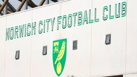 Green writing saying "Norwich City Football Club" on a white external wall of the Carrow Road stadium above the club's logo of yellow canary on a green shield.