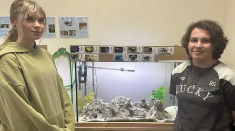 Brooke Chaplow and Kall Stott stand in front of a fish tank. Brooke has blonde hair and wears a lime green hoodie while Kall has cropped brown hair and a T-shirt that reads 'LUCKY'. The fish tank has pictures of fish stuck to the top of it.