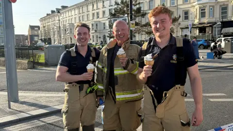 Jerome's Pavillion/Harrison Leisure Three male firefighters wearing beige uniform trousers and navy polo shirts eat ice cream cones in the street.