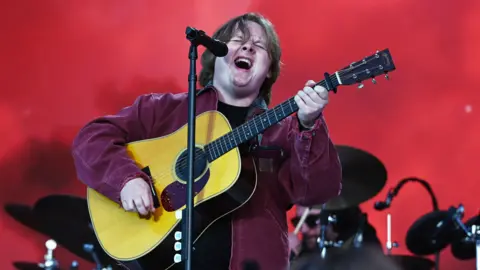 BBC/Sarah Jeynes/Jamie Simonds Lewis Capaldi wearing a burgundy coat and holding a guitar while singing into a microphone on stage.