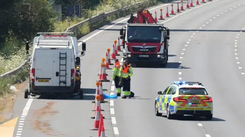 A close-up view of a motorway lane shows orange traffic cones stretching into the distance. A marked police car with blue lights, a white recovery van and a red recovery lorry are parked in the closed lanes. Two highway workers wearing hi-vis clothing stand beside the cones.