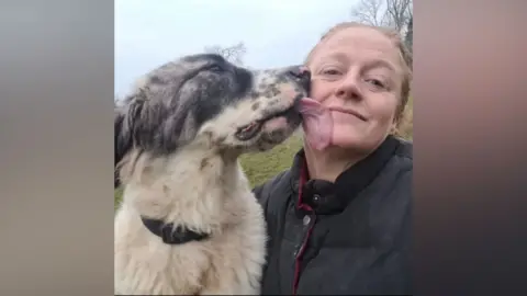 Teresa Underwood A close-up outdoor selfie showing a woman wearing a dark jacket while a large, fluffy dog with grey and white fur reaches up and licks the person’s cheek. Grass and trees are visible in the background.