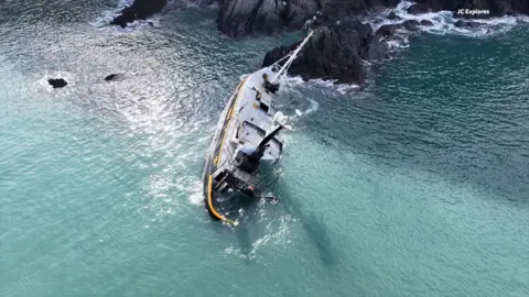 Wide drone shot shows the large navy and orange vessel sunk in the sea. The boat is leaning on one side as the front of the boat is trapped by black rocks and is submerged in blue water.