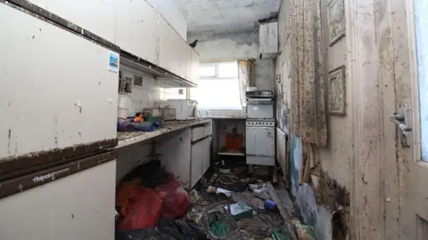 Blackpool Council The galley kitchen with white units to the left and a wall with a curtain to the right and at the far end. There is rubbish on covering the dirty floor 