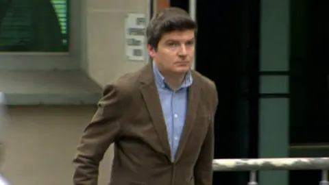 Michael McMonagle - A man with short dark hair, a collared blue shirt and a brown suit jacket. He is walking outside of a white bricked building.