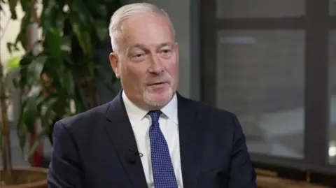 Richard Fuller, with short grey hair, is seated indoors during an interview, wearing a dark suit, white shirt and blue patterned tie. The background is softly blurred, showing a large green plant and office-style windows. A lapel microphone is clipped to the jacket.