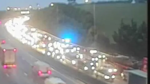 A still taken from a traffic camera looking at the M25. Vehicles are queuing across four lanes on the anti-clockwise carriageway. The blue lights of an emergency vehicle can be seen among the congestion.