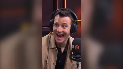 Luke Thompson has a big smile. He has short brown hair and wears a camel-coloured jacket and black headphones