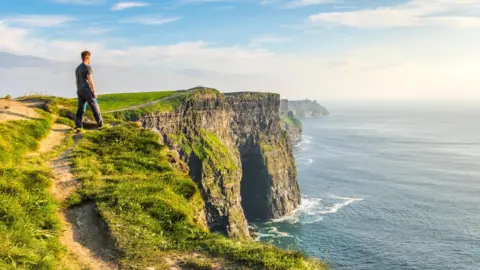 A wide angle shot cliffs by the ocean, there is a man looking off into the horizon