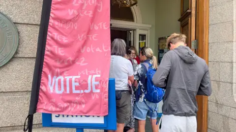 People queue outside a polling station in Jersey. There is a Vote.je sign by the doorway.