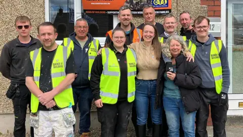 Vicky with her mum and some of the volunteers, many wearing yellow hi-viz tabards over their work gear, outside the house.
