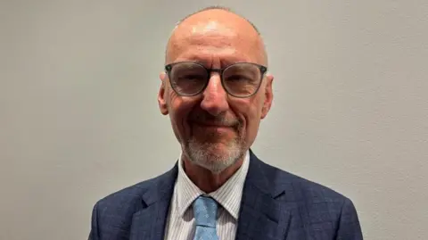 The image shows a man standing against a plain, light grey wall, facing the camera directly. He has a bald head and is wearing round, dark-framed glasses. He has a trimmed moustache and beard around the chin area. He is wearing a blue textured suit jacket over a light-coloured shirt with narrow vertical stripes. A light blue tie with a small repeating pattern is tied at the collar.