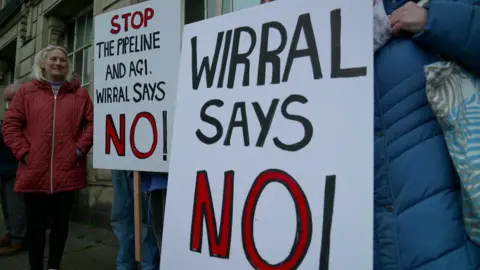 Protesters outside Wallasey Town Hall hold up signs opposing the Peak Cluster scheme which read: 'Wirral Says No' and 'Stop the pipeline and AGI Wirral Says No'.