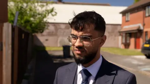 A man in a navy suit with pale blue check pattern, white shirt and navy tie, stands in a suburban street with houses in the background. he has a dark brown beard and hair with a fringe. He is wearing silver wire-framed glasses and looking to the right of the camera at something and is pictured on a sunny day.