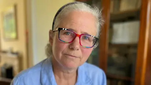 Heather is wearing a blue blouse and brightly-framed glasses. Her hair is tied back from her face and she has a neutral expression. She is in her living room with bookshelves and a television screen in the background, out of focus.