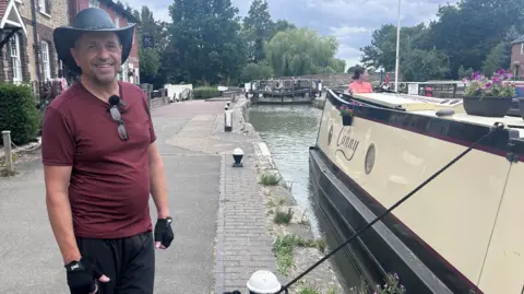 A man, wearing a hat, red T-shirt, black trousers and gloves is standing on a path next to a canal boat, smiling. A building is behind him and there is a lock in the distance. 