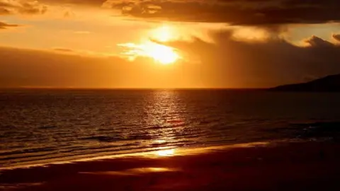 Graham Christie a golden sunset over a calm sea, with dark clouds drifting across the sky and warm light reflecting on gentle waves along the shoreline.