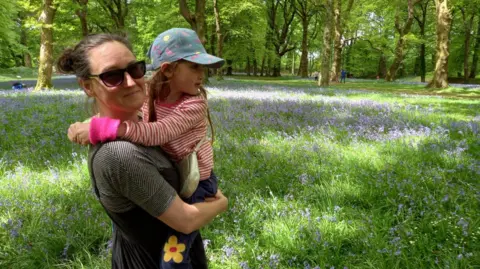 A woman, wearing dungarees and a striped top. She is turned to the side and is looking at the camera. She is wearing sunglasses. In her arms is a young child in a red and white striped top and blue cap and facing to the right. 
Behind them is grass and purple flowers. There are trees in the background.