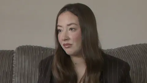 A woman with long brown hair, wearing a black blazer, photographed from the chest up while sitting on abrown sofa