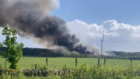 A fire next to a wind turbine in a green field with clouds of black smoke billowing into the sky 