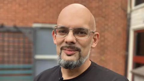 Luke Dadswell an advanced support worker, he is wearing a black T-shirt, standing outside the Gilmorton supported living service flats in Leicester 