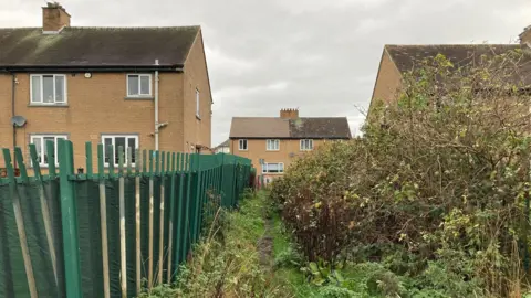 A ginnel flanked by a green metal fence going in between two semi-detatched brown brick houses