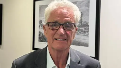 A man wearing a grey suit adn white shirt. He has black glasses and grey hair and is stand in front of a black and white photo in a frame.