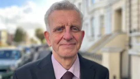 Councillor Trevor Muten is wearing a suit and is standing on the pavement of a busy road in Hove, with a number of parked cars behind him. 