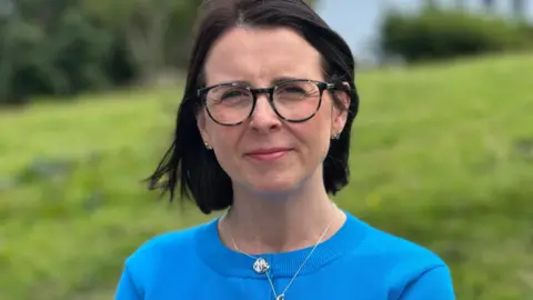 Sarah Maltby, who has shoulder-length dark hair and is wearing dark rimmed glasses and a blue round-necked top. There is a green grass slope behind her.