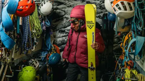 Jan Virt Fay Manners, holding her skis, in a room full of ropes and other hardware made for climbing. A print of a snowy mountain range is fixed to the wall.
