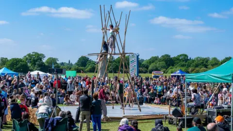 Nene Park Trust An acrobatic performance of two people standing on bamboo sticks with people watching them.