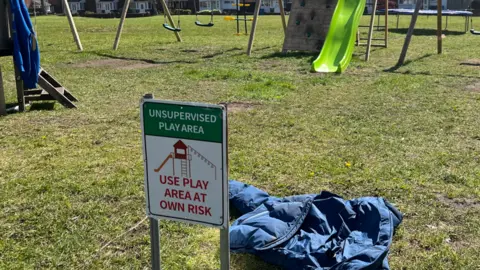 Jonny Humphries/BBC A small sign, posted into a grassy field, reads 'Unsupervised Play Area - Use Play Area At Own Risk'. In the back ground are two sets of swings and a green slide next to a wooden board with climbing-wall style hand and foot holds bolted onto it. 