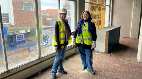 Clare Worden/BBC BBC reporter Clare Worden stands on the left of the image in high-vis and hard hat. She is wearing jeans and black sweatshirt. She is holding a microphone towards Morgan Ellis Leah who is wearing a high-vis vest, blue scarf and jeans. They are standing inside a derelict building by its large windows.