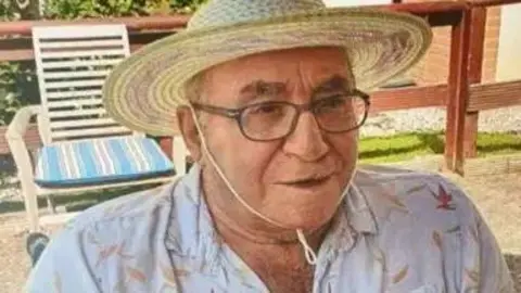 An elderly man wearing glasses and a sun hat, while wearing a white shirt. He is sat in a chair outside looking off to the right of the camera