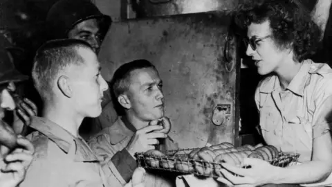 Getty Images Two American soldiers, who are brothers, sample doughnuts offered by a member of an American Red Cross Clubmobile in Normandy (summer 1944). She is on the right with the tray in front of her. The two men are on the left and looking up at her, holding a doughnut each. 