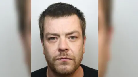 A mug shot of Stuart Hand who is a convicted paedophile. He is a white man with short brown hair and green eyes. He is wearing a black t-shirt. 