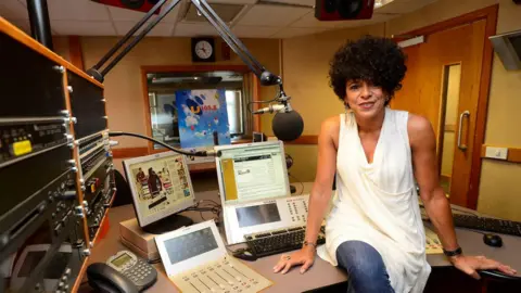 Pacemaker A woman with curly dark hair and dangly earrings sits on a radio production desk. She wears a white sleeveless top and blue jeans. Behind her is radio production technology iand computer screens.