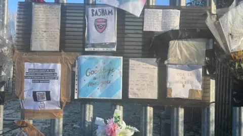 Tributes, cards and flowers attached to silver railings. One card says "Goodbye, we'll miss you" and another says "You'll Be Missed." There are flowers at the bottom of the picture and to the right.
