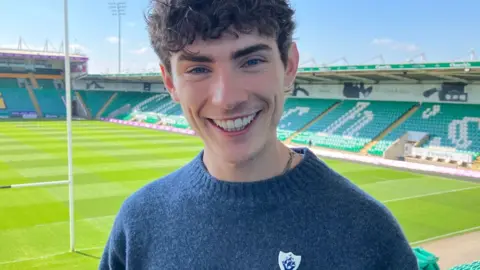 Sam Read/BBC Joel Mawhinney, who has short curly hair, is wearing a blue jumper with a Blue Peter badge pinned to it and is smiling broadly. Behind him can be seen Northampton Saints' stadium, pitch and stands