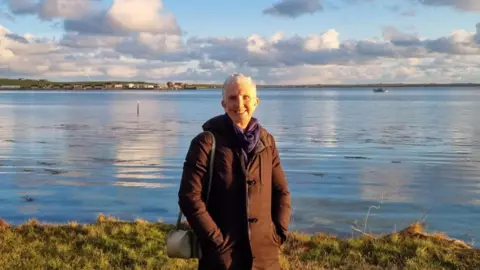 Stewart Bain Ann Cleeves, wearing a brown jacket and purple scarf. She is smiling and standing in front of a body of water.