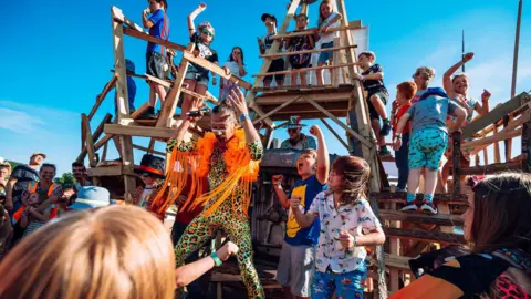 A man in a green and orange leopard print catsuit with an orange shawl and children dancing and singing next to him. There is a wooden climbing stand behind them with children climbing on it and a DJ at a booth within the wooden construction. There is a crowd of people to the sides and behind the camera and the sky is bright blue.