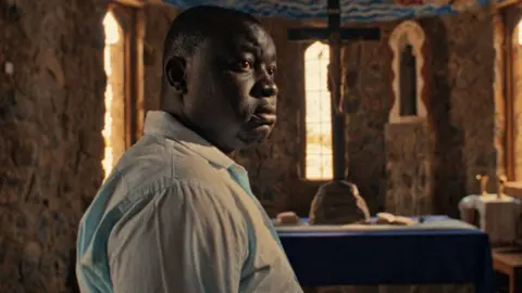 FILMCREW Ben Tekee as the character Shadrack, wearing a pale blue shirt, in the film Nawi. He is seen in a church - a giant cross seen on an small alter in the background